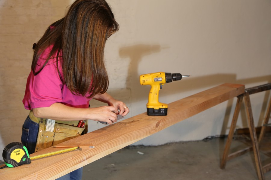 A woman wearing a toolbelt, looking down on a plank of wood with a tape measure and drill on it